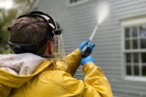 Person cleaning house siding with a pressure washer, wearing protective gear in a yellow raincoat.
